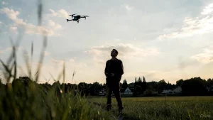 Mann fliegt Drohne auf einem Feld mit sicherem Abstand zu Wohnhäusern im Hintergrund.